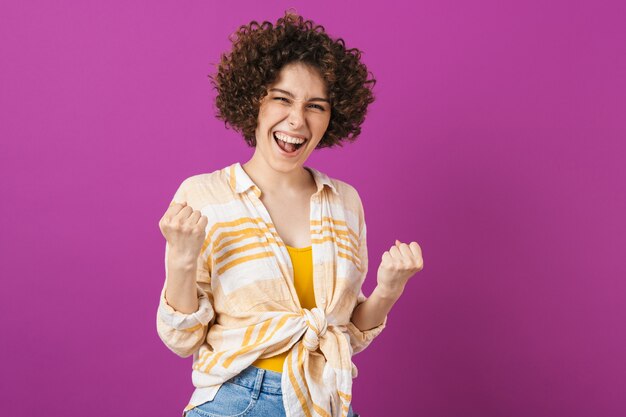 Retrato de una atractiva joven alegre con cabello rizado morena que se encuentran aisladas sobre la pared violeta, celebrando el éxito