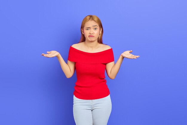 Retrato de una atractiva joven asiática confundida con el pelo rubio corto vestido rojo de pie y encogiéndose de hombros aislada sobre un fondo morado