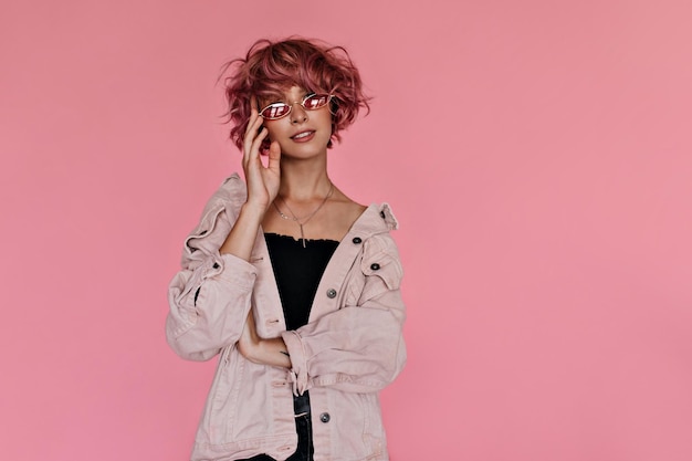 Retrato de una atractiva joven de cabello rosado con gafas de sol rojas Una chica rizada fresca con una chaqueta de mezclilla de gran tamaño posa sobre un fondo rosa aislado