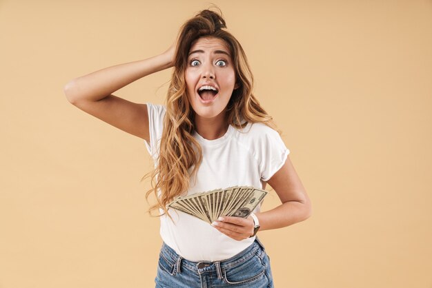 Foto retrato de una atractiva joven emocionada con largo cabello rubio rizado vistiendo ropa casual que se encuentran aisladas, mostrando billetes de dinero