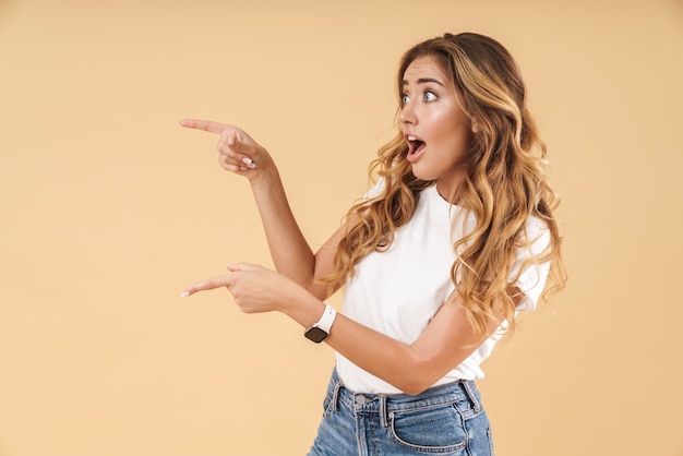 Foto retrato de una atractiva joven emocionada con largo cabello rubio rizado vistiendo ropa casual que se encuentran aisladas, señalando con el dedo en el espacio de la copia