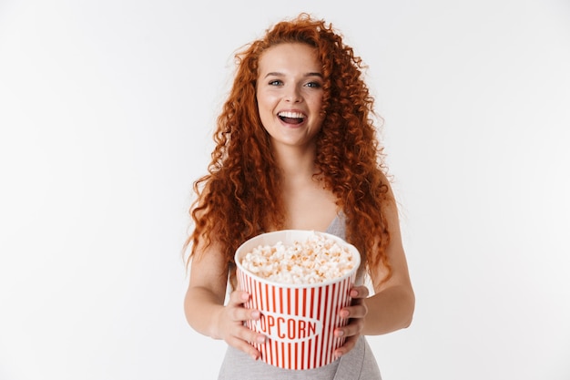 Retrato de una atractiva joven emocionada con el pelo rojo largo y rizado que se encuentran aisladas, viendo una película y comiendo palomitas de maíz