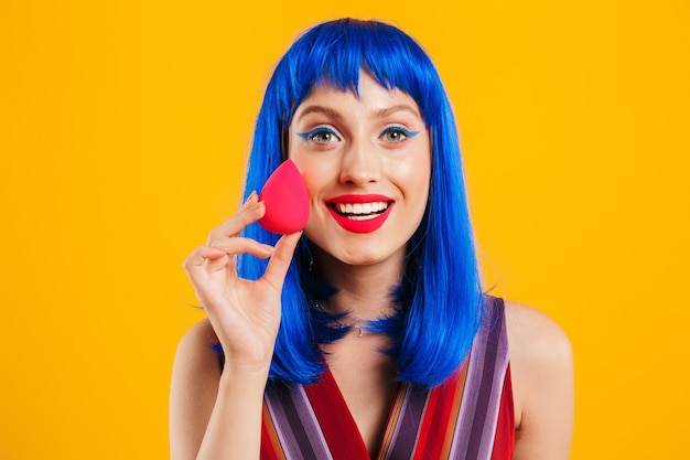 Retrato de una atractiva joven funky sonriente con cabello azul vistiendo ropa casual que se encuentran aisladas sobre la pared amarilla, mostrando la esponja de maquillaje,