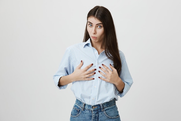 Retrato de una atractiva modelo femenina con expresión impresionada sosteniendo su pecho con ambas manos y levantando las cejas mientras estaba de pie sobre un fondo gris La mujer compró un nuevo sostén pushup y quedó satisfecha