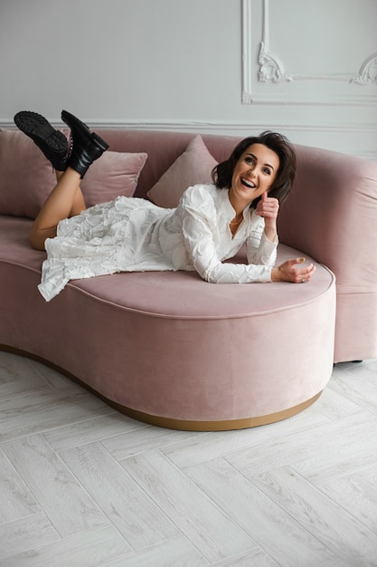 Retrato de atractiva morena alegre en vestido blanco y botas de cuero negro tendido en el sofá de terciopelo rosa con cojines sonriendo y mirando a la cámara.