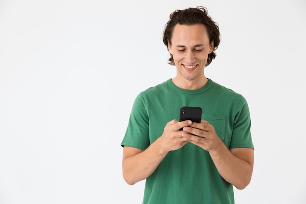 Retrato de una atractiva morena hombre vestido con camiseta básica sonriendo y sosteniendo el teléfono inteligente aislado sobre la pared blanca