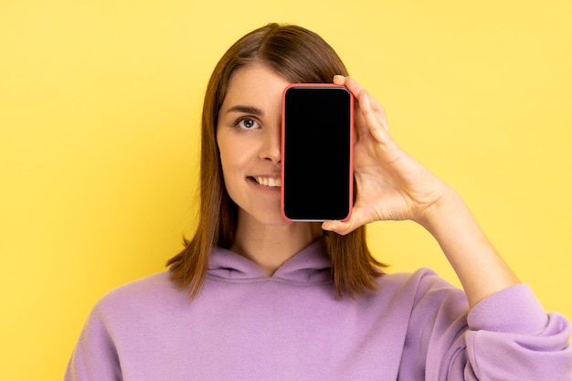 Retrato de una atractiva mujer adulta joven con cabello oscuro que cubre los ojos con un teléfono celular con pantalla vacía, con capucha morada. Disparo de estudio interior aislado sobre fondo amarillo.