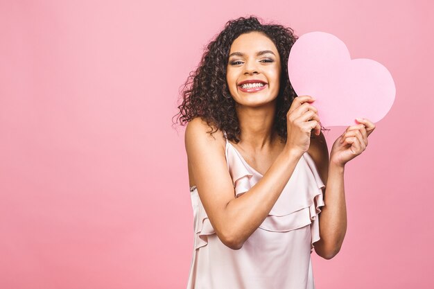 Retrato de atractiva mujer afro americana sonriente feliz aislada sobre fondo rosa con gran corazón rosa