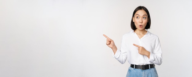 Retrato de una atractiva mujer asiática adulta que señala mirando a la izquierda con una sonrisa complacida que muestra una pancarta o un logotipo a un lado de pie contra el fondo blanco del estudio