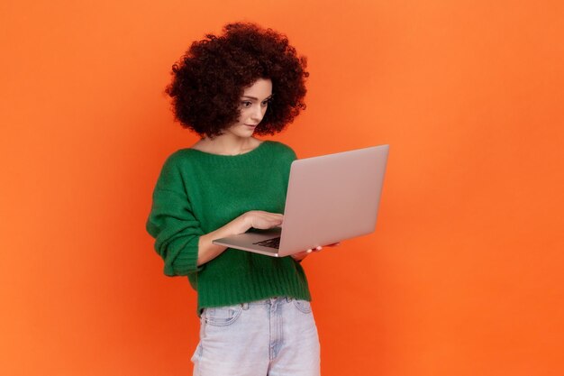 Retrato de una atractiva mujer concentrada con peinado afro que usa un suéter de estilo informal verde que sostiene un portátil independiente que trabaja en línea Foto de estudio interior aislada en un fondo naranja