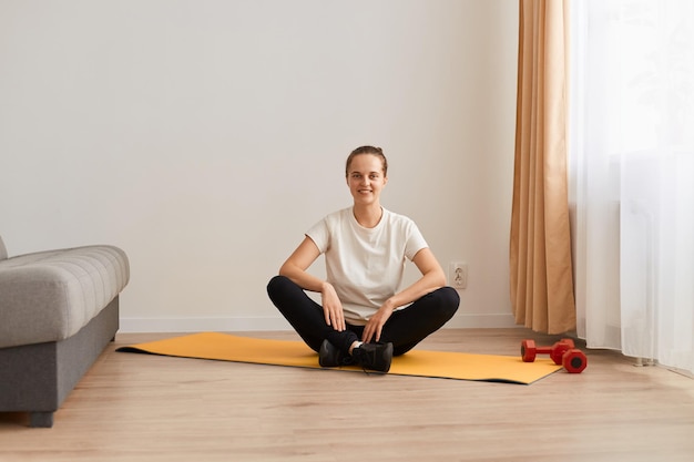 Retrato de una atractiva mujer deportiva sentada en una pose de loto en una alfombra, usando una camiseta blanca y leggins negros, mirando la cámara con una sonrisa feliz, meditando y entrenando en casa en la sala de estar.