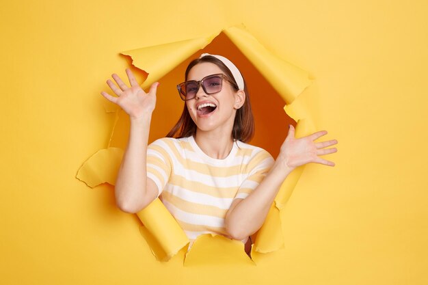 Retrato de una atractiva mujer feliz con camisa a rayas y banda para el pelo posando en un agujero de papel amarillo de pie con los brazos levantados celebrando su victoria expresando emociones positivas