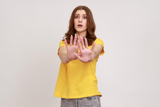 Retrato de una atractiva mujer insatisfecha con el pelo castaño ondulado frunciendo el ceño mostrando un gesto de parada con ambas manos usando una camiseta amarilla casual Foto de estudio interior aislada en fondo gris