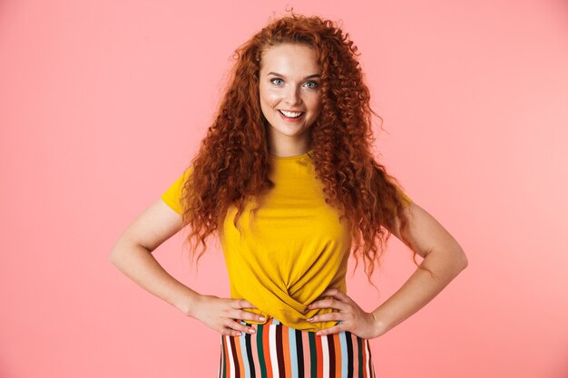 Foto retrato de una atractiva mujer joven feliz con el pelo rojo largo y rizado que se encuentran aisladas, con las manos en las caderas