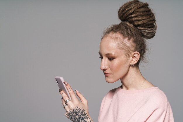 Retrato de una atractiva mujer joven con rastas que se encuentran aisladas sobre la pared gris, sosteniendo el teléfono móvil