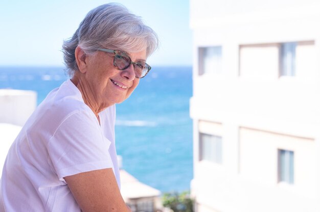 Retrato de una atractiva mujer madura relajante en el balcón de su casa, contemplando el mar y el cielo azul durante un día soleado al aire libre.