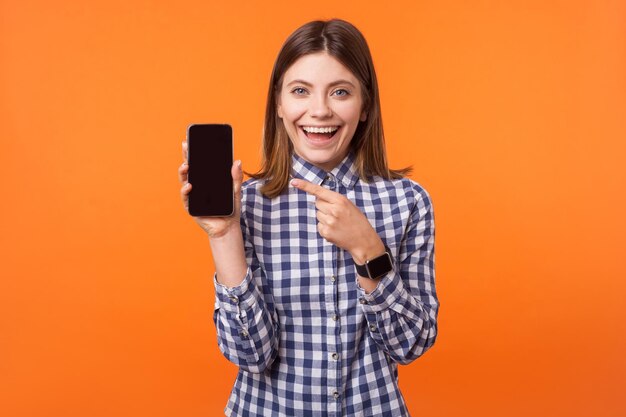 Retrato de una atractiva mujer morena con una sonrisa encantadora con una camisa a cuadros de pie apuntando al teléfono satisfecha con aplicaciones y servicios móviles en un estudio interior aislado en un fondo naranja