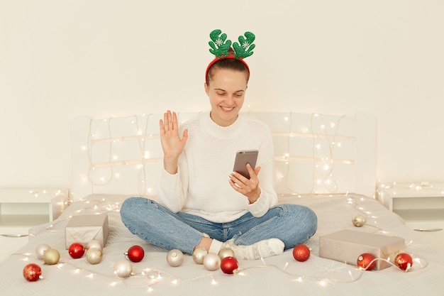 Retrato de atractiva mujer positiva vestida con jeans suéter blanco y aro de accesorios de fiesta, sentada en la cama en el dormitorio, teniendo videollamadas a través del teléfono móvil, saludando con la mano, felicitando con la Navidad.