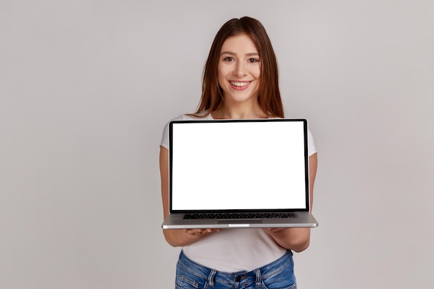 Retrato de una atractiva mujer sonriente sostiene una computadora portátil con una pantalla blanca para la promoción del sitio web del servicio en línea de publicidad en Internet con una camiseta blanca Foto de estudio interior aislada en un fondo gris