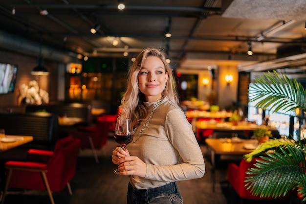 Retrato de una atractiva mujer sosteniendo una copa de vino tinto parada en un restaurante con un interior de lujo con un interior oscuro mirando hacia otro lado