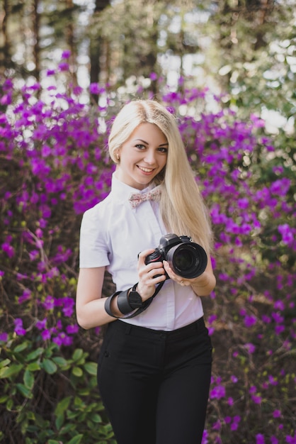 Retrato de una atractiva rubia sonriente mujer fotógrafa en una camisa con una cámara en sus manos contra flores de color púrpura