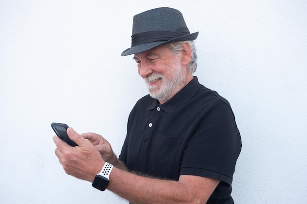 Retrato de un atractivo anciano barbudo de negro con sombrero usando un teléfono móvil para enviar mensajes aislado de fondo blanco