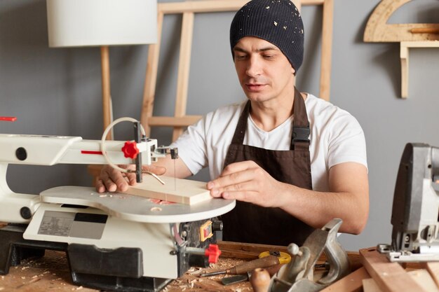 Retrato de un atractivo carpintero caucásico con delantal marrón y gorra negra usando una sierra eléctrica trabajando en su carpintería disfrutando del proceso de fabricación de productos de madera