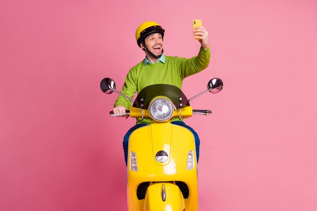 Retrato de atractivo chico alegre montando moto tomando selfie en teléfono