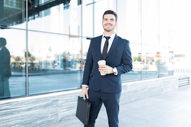 Retrato de un atractivo director ejecutivo masculino con una taza de café para llevar y un maletín parado frente al edificio de oficinas