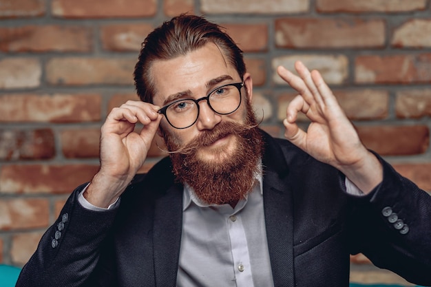 Retrato de un atractivo hombre brillante y elegante con gafas, con bigote y barba, mostrando bien con los dedos.