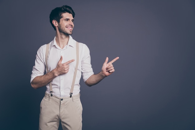 retrato de atractivo hombre de negocios macho indicando los dedos espacio vacío buenas noticias oferta vestido ropa formal camisa blanca pantalones de tirantes beige.