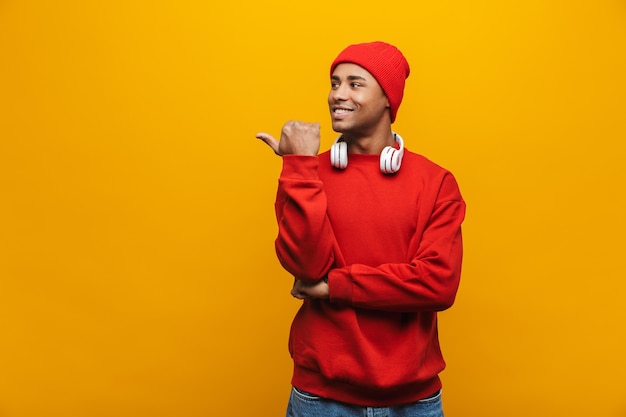 Retrato de un atractivo joven africano casual confiado sonriente de pie sobre la pared amarilla, apuntando con el dedo hacia el espacio de copia