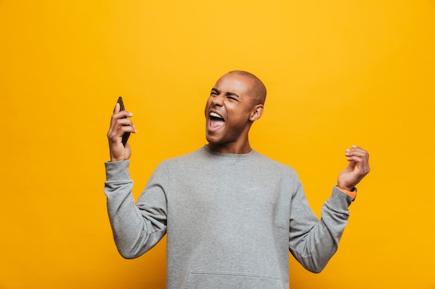 Retrato de un atractivo joven africano casual enojado de pie sobre la pared amarilla, sosteniendo el teléfono móvil