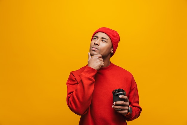 Retrato de un atractivo joven africano casual pensativo de pie sobre la pared amarilla, sosteniendo la taza de café para llevar