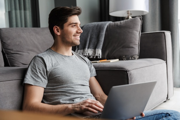 Retrato de un atractivo joven barbudo sonriente vistiendo ropa casual sentado en un piso en el sofá de la sala de estar, usando la computadora portátil