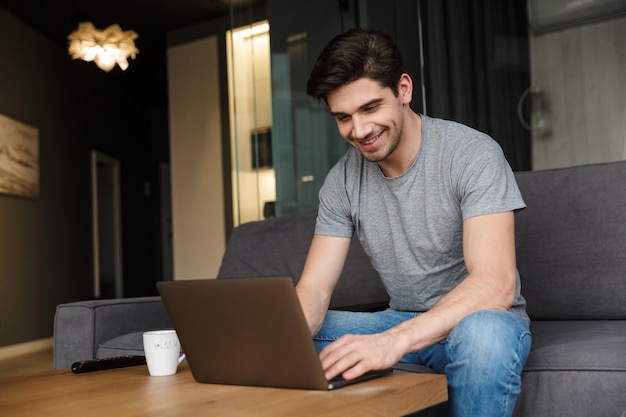 Retrato de un atractivo joven barbudo sonriente vistiendo ropa casual sentado en un sofá en la sala de estar, usando la computadora portátil