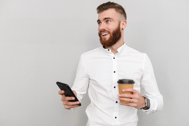 Retrato de un atractivo joven empresario de pie sobre una pared gris, mediante teléfono móvil mientras bebe café para llevar