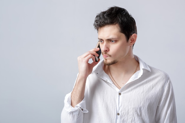 Retrato de atractivo joven morena con camisa blanca hablando por el teléfono inteligente sobre el fondo gris.