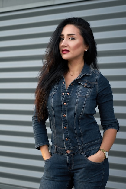 Retrato atractivo joven morena de moda con el pelo largo y labios rojos con overoles de mezclilla moderna sobre fondo gris pared urbana.