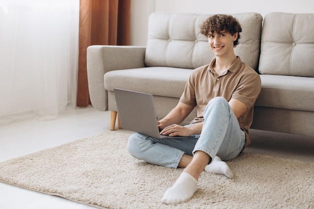 Retrato de un atractivo joven de pelo rizado que usa una laptop mientras se sienta en el sofá en casa