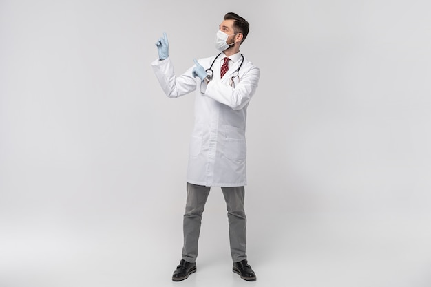 Retrato de atractivo médico guapo en mascarilla protectora, bata de laboratorio blanca, corbata aislada en la pared blanca