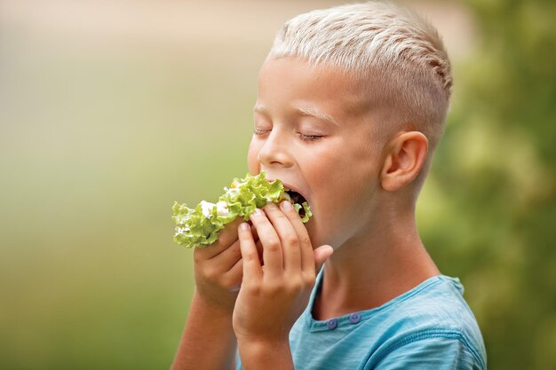 Retrato de un atractivo niño caucásico comiendo ensalada en un día soleado de verano en el parque enfocado en una boca con ensalada