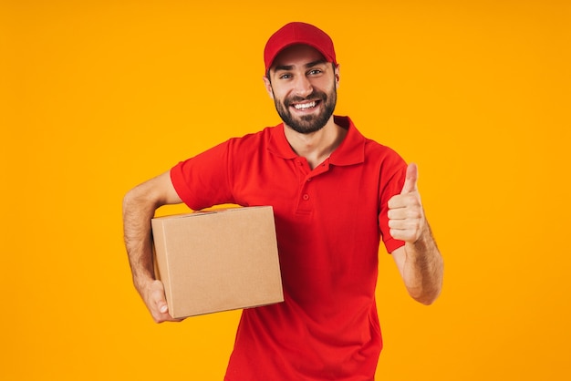 Retrato de atractivo repartidor en uniforme rojo mostrando el pulgar hacia arriba y sosteniendo la caja de embalaje aislada sobre amarillo