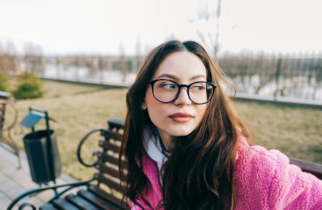 Retrato autêntico de uma jovem mulher caucasiana com cabelo morena em óculos, sentado em um banco no parque.