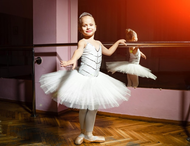 Retrato de bailarina joven sonriente en tutú blanco y pointes posando aislada en el estudio de danza Pequeña bailarina de balet de pie cerca del bar y el espejo preparándose para la actuación