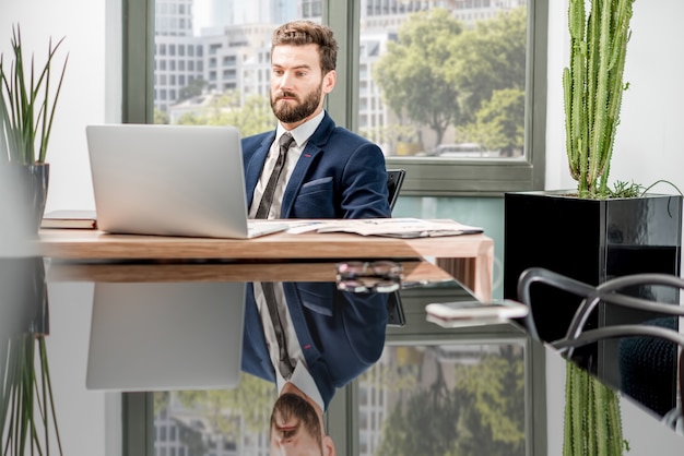 Retrato de un banquero guapo que trabaja con la computadora portátil sentado en el interior de la oficina de lujo con hermosas vistas a los rascacielos