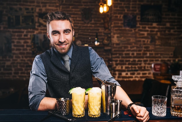 Retrato de barman guapo profesional sonriendo a la cámara preparando cócteles y sirviendo bebidas frescas