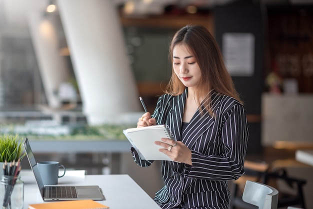 Retrato de bastante joven empresaria asiática tomando nota y usando la computadora portátil en la oficina moderna