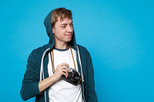 Retrato de bastante joven en ropa casual mirando a un lado, sosteniendo la cámara de fotos vintage retro aislada en la pared azul. Personas sinceras emociones, concepto de estilo de vida.