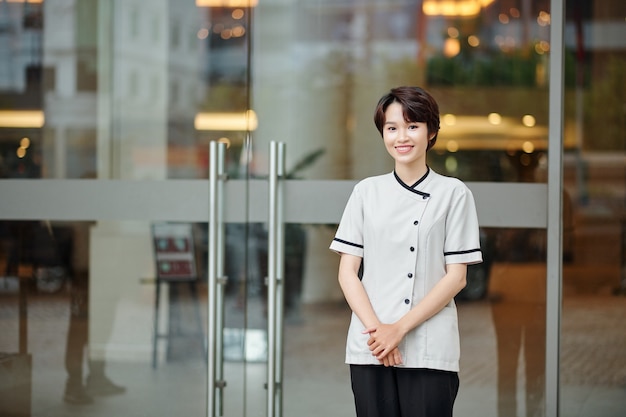 Retrato de bastante joven trabajador hotelero vietnamita de pie en las puertas de cristal del hotel y sonriendo a la cámara